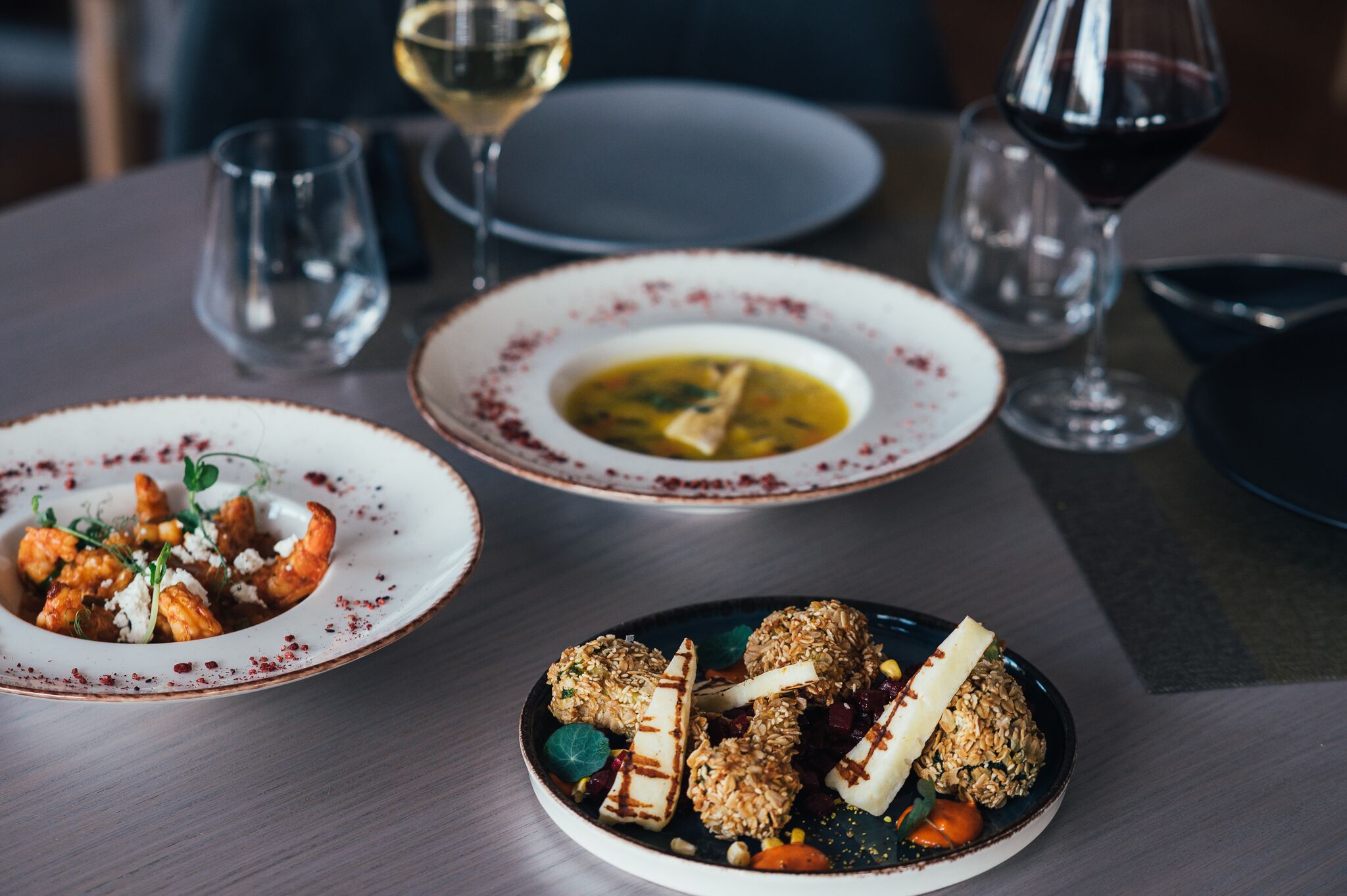 A dining table with three plates of food, two glasses of wine, and a bowl.