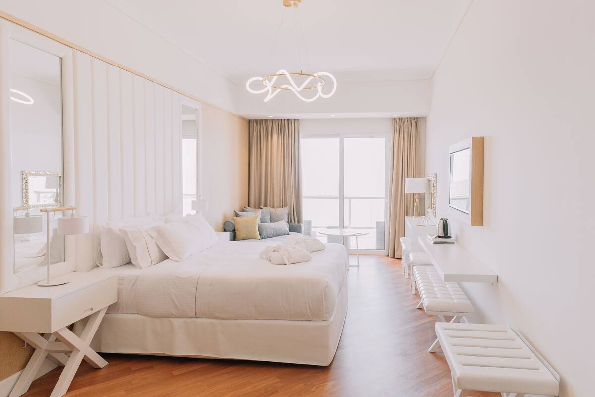 A modern hotel room with a large bed, a vanity with a mirror, a desk, and a view of the city through large windows.