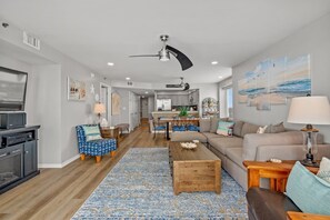 Un salon spacieux avec une table basse en bois, un canapé et un téléviseur. Au fond, on aperçoit une cuisine avec un coin repas.
