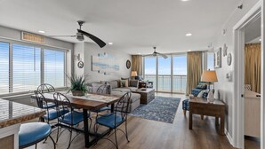 Een ruime woonkamer met een eettafel, blauwe stoelen, een bank en een balkon met uitzicht op zee.