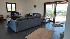 A modern living room with a grey sofa, a wooden coffee table, and a TV on a stand. There is a large window with a view of the countryside. - Villa Las Adelfas with beautiful view ,HEATED POOL, 6 Pers, WIFI,BBQ,terraces (Benamargosa)