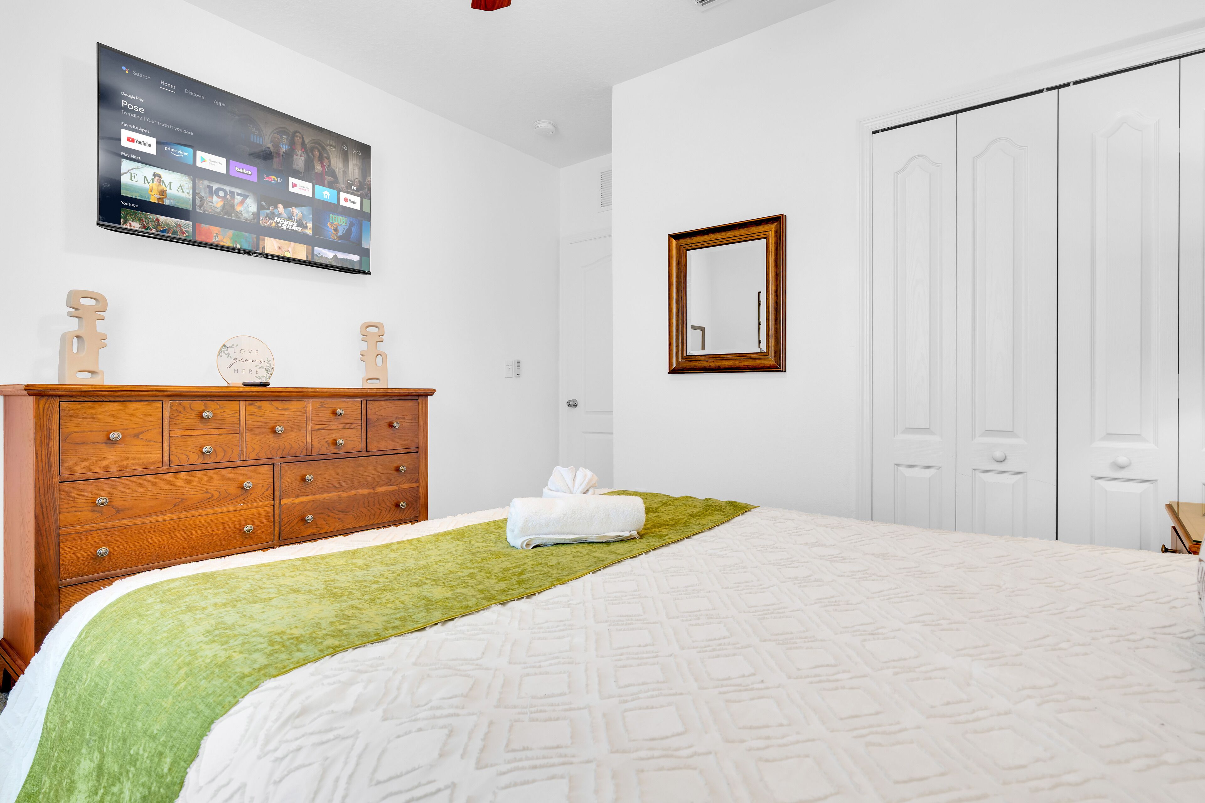 A bedroom with a bed, a wooden dresser, a television mounted on the wall, and a wardrobe.