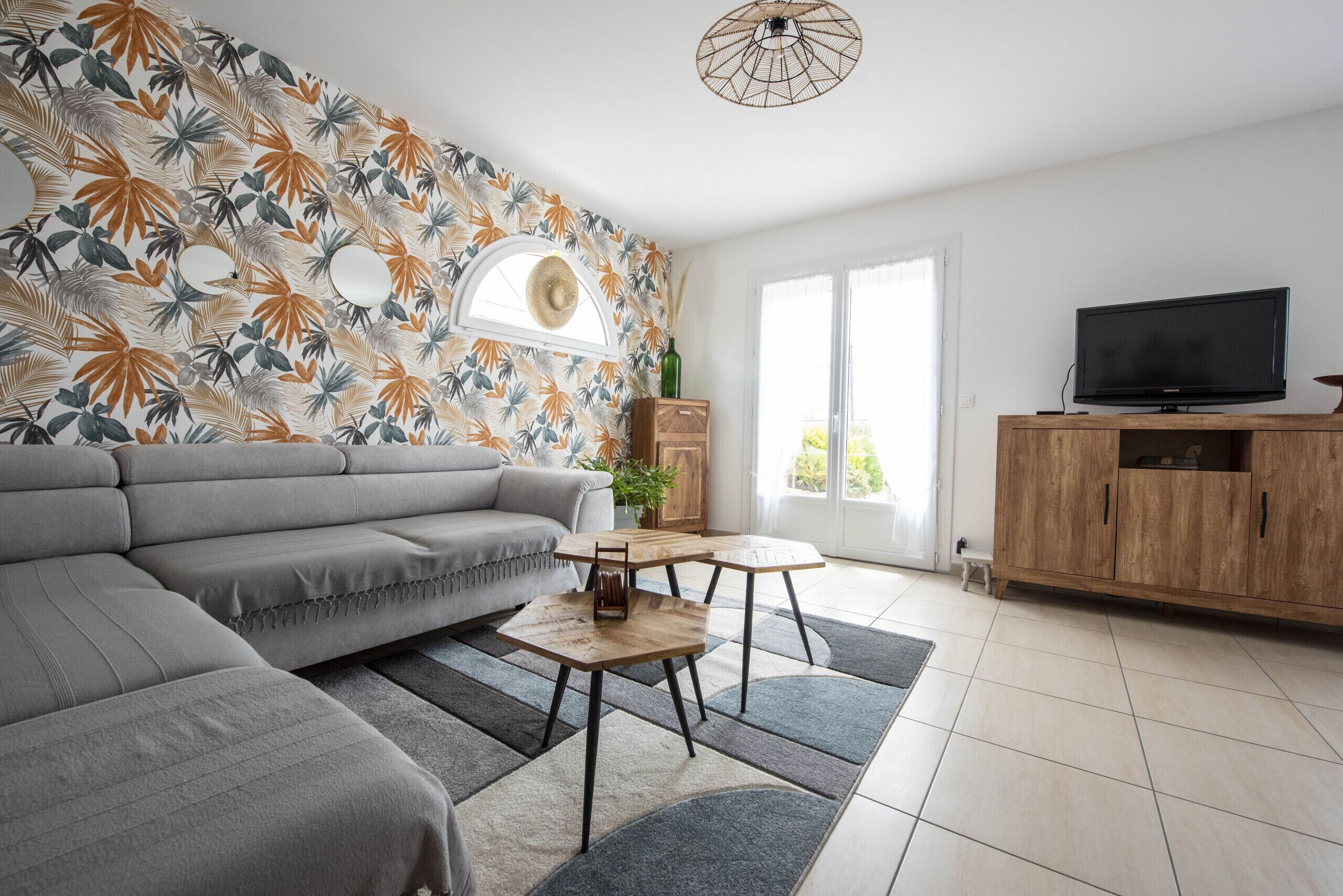 Un salon moderne avec un canapé gris, deux tables en bois, un téléviseur posé sur un meuble en bois et un mur orné d’un motif de feuilles tropicales.