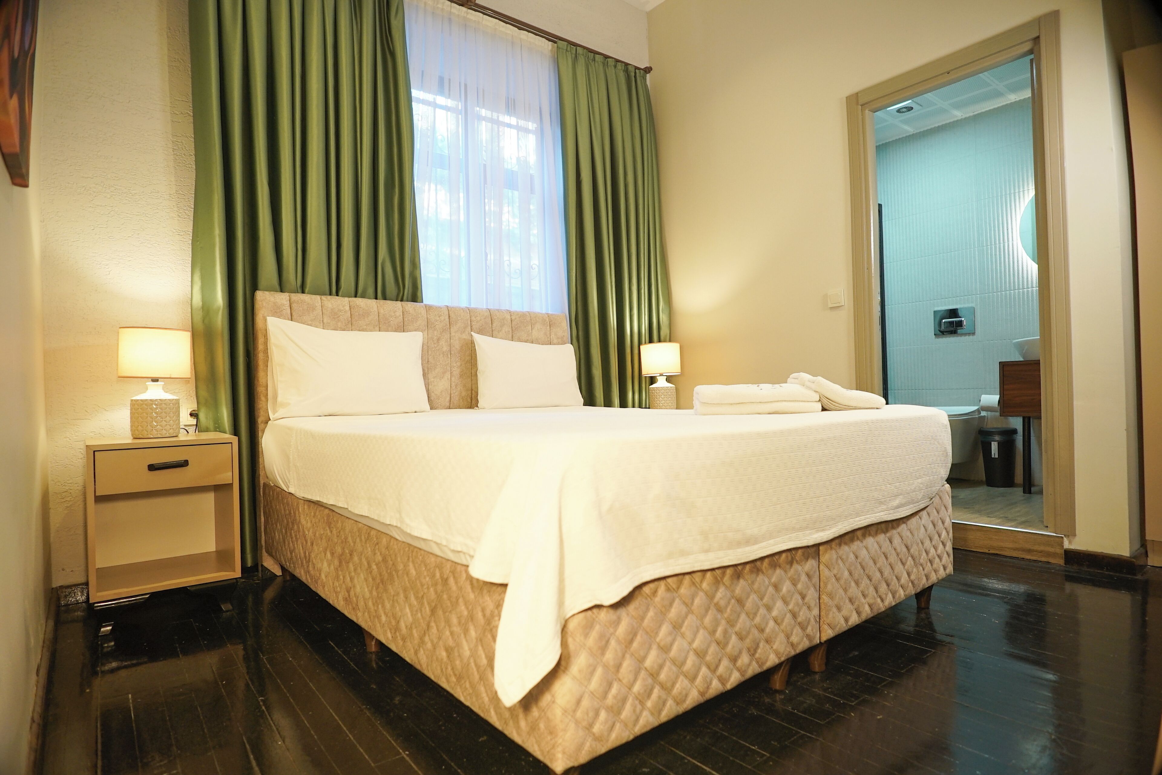 A neatly made bed with white linens, a nightstand with a lamp, and a mirror in the adjacent room.