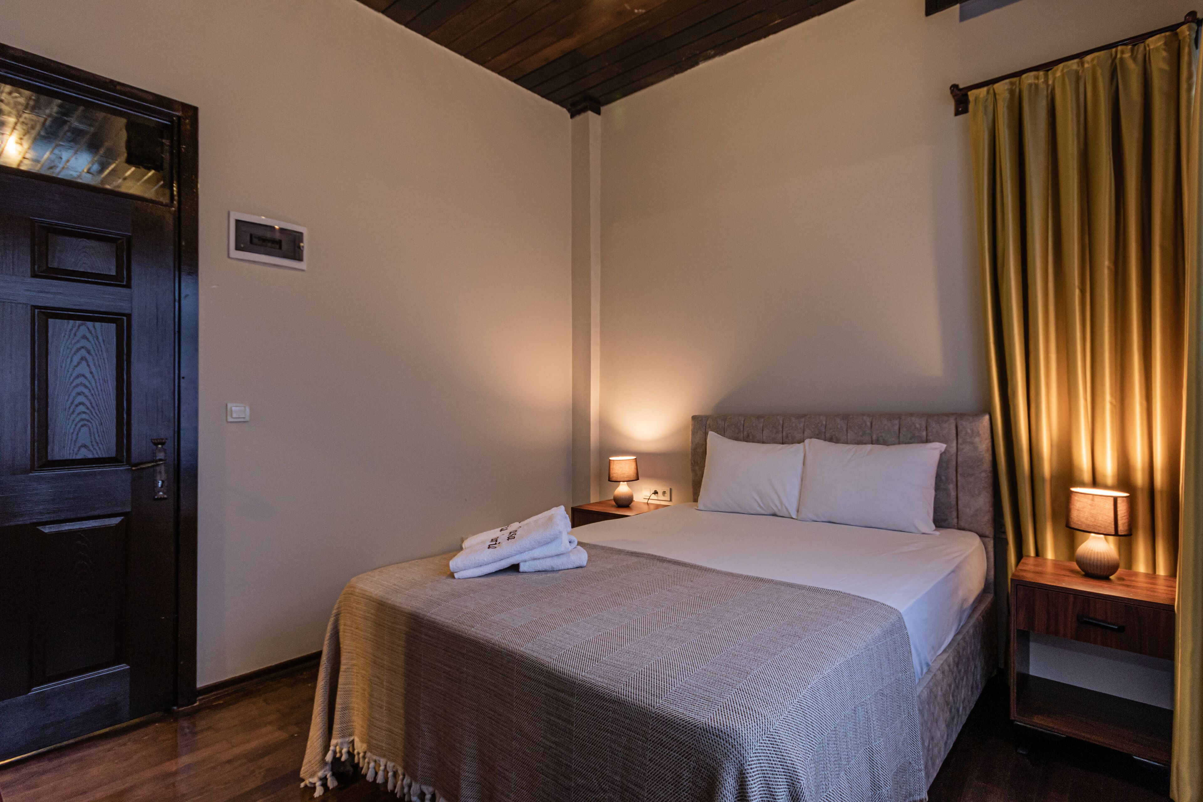 Ein ordentlich bezogenes Bett mit weißer Decke und Kissen, eine Holztür, ein Nachttisch mit Lampe und ein Vorhang.