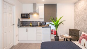 A compact kitchen with white cabinets, a microwave, and a small refrigerator. There's a potted plant and a dining area with a chair and table.