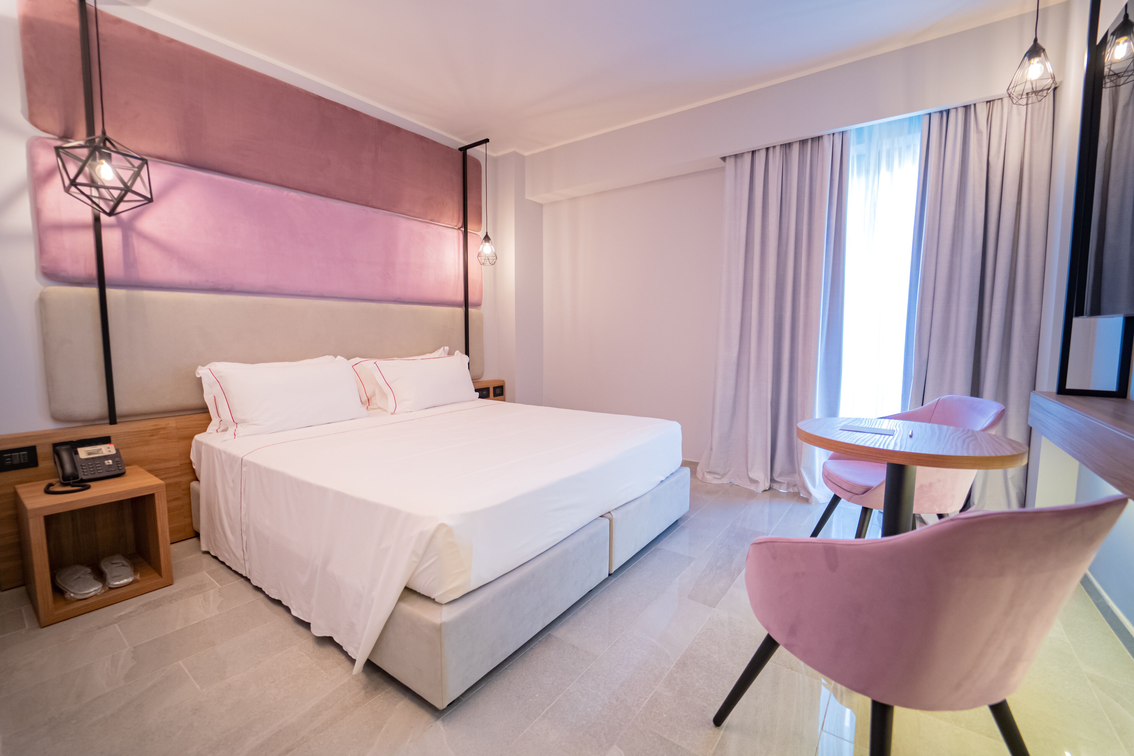 A modern hotel room with a large bed, bedside table with a phone, a small table with a chair, and a view of the outside through the window.