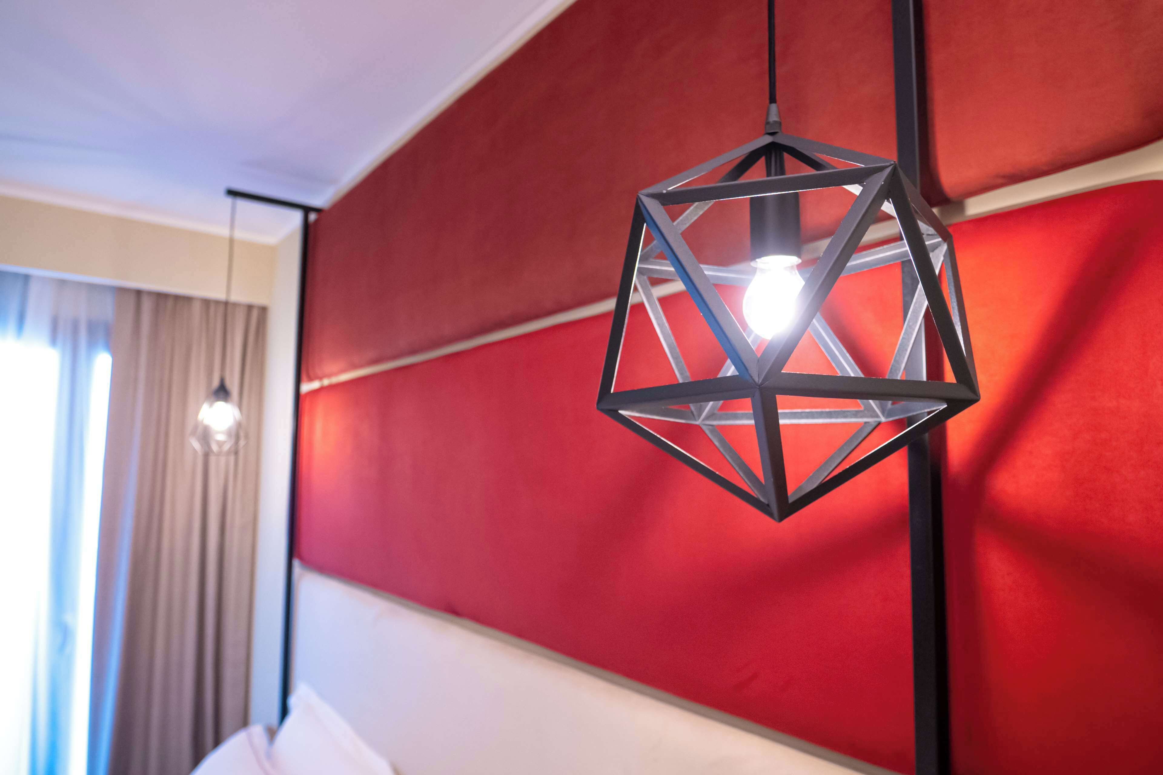 Een geometrische hanglamp met zichtbare gloeilamp, hangend aan een zwarte metalen standaard, in een kamer met een rode accentmuur en een witte plint.