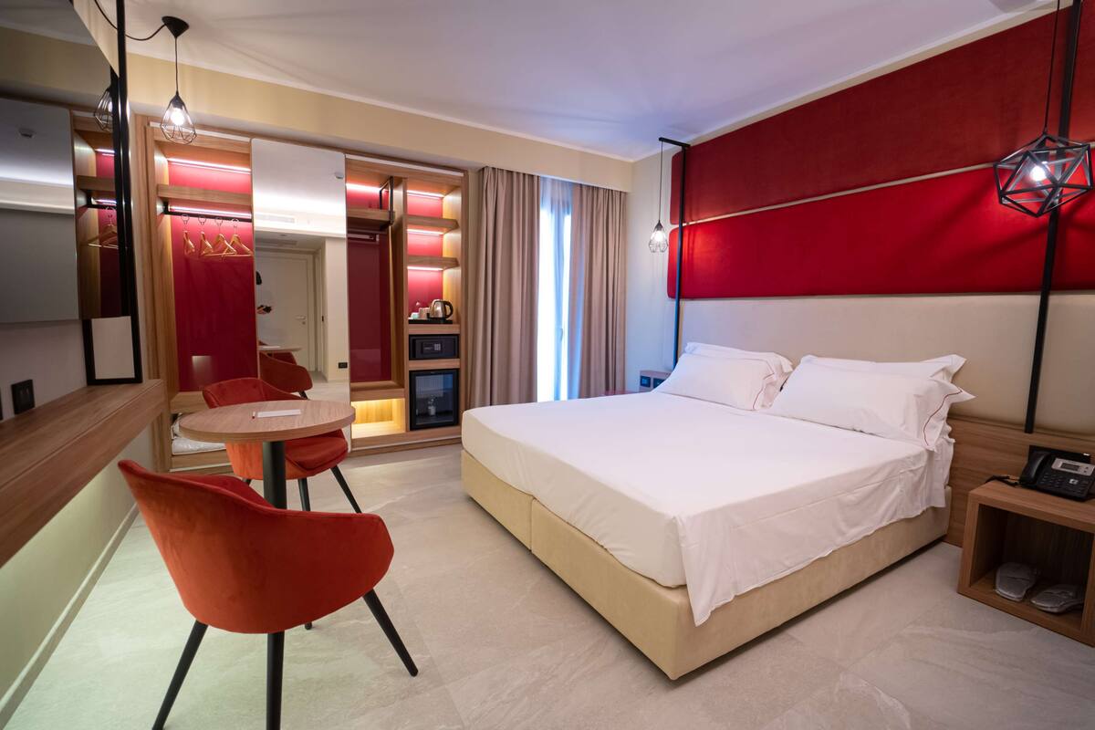 A hotel room with a bed, a desk with a chair, a wardrobe, and a bedside table with a phone.