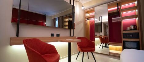 A modern hotel room with a round table, two red chairs, a large mirror, and a built-in wardrobe with red lighting.