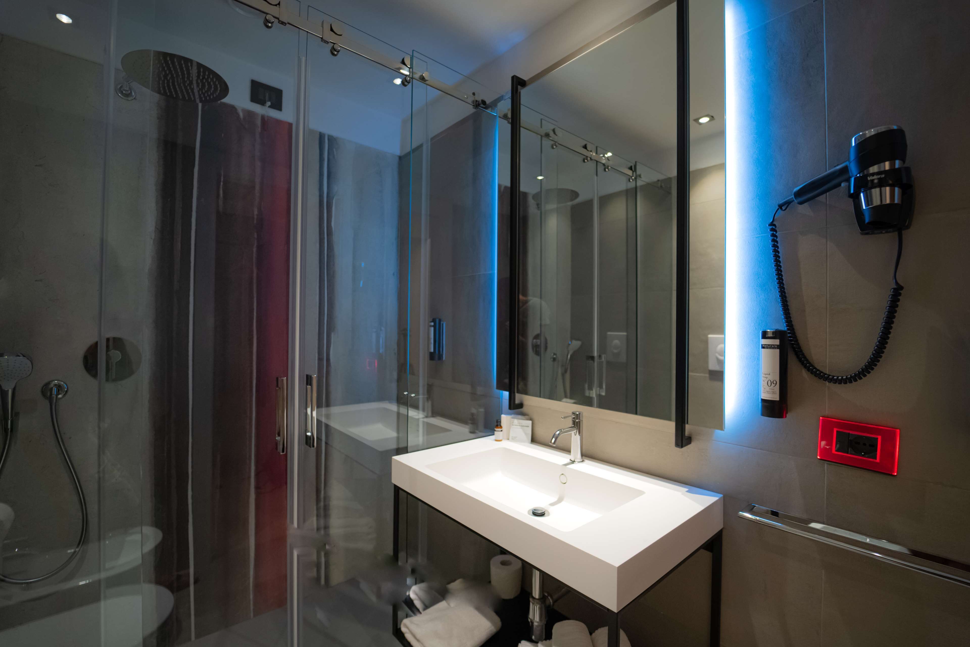 A modern bathroom with a glass shower enclosure, a white sink, and a hairdryer mounted on the wall.