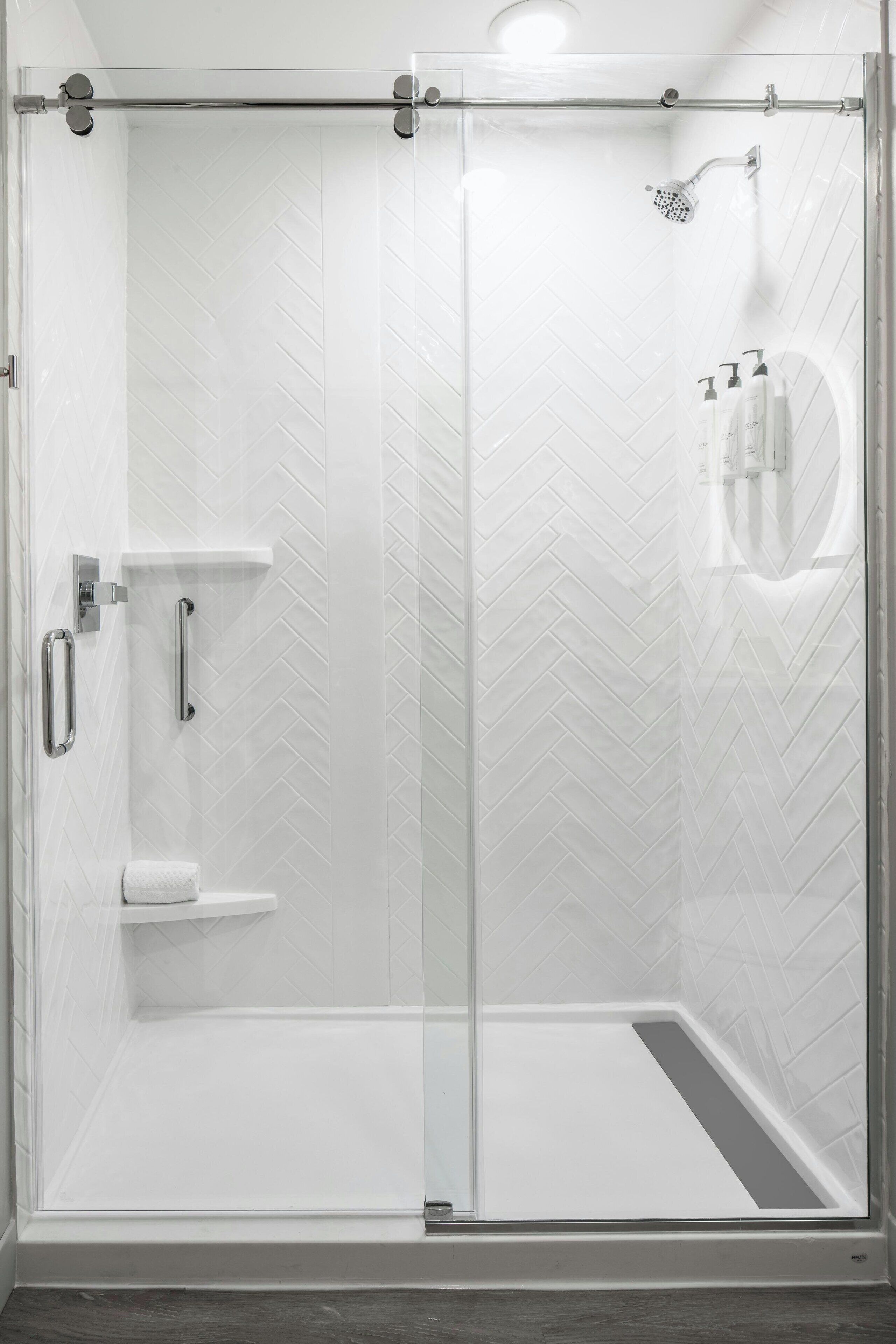 A glass-enclosed shower with a white herringbone tile pattern, a handheld showerhead, and a small shelf with toiletries.