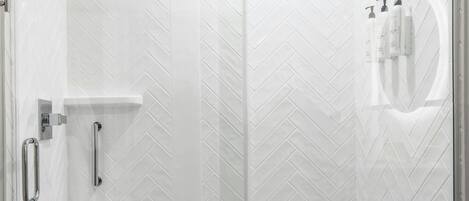 A glass-enclosed shower with a white herringbone tile pattern, a handheld showerhead, and a small shelf with toiletries.