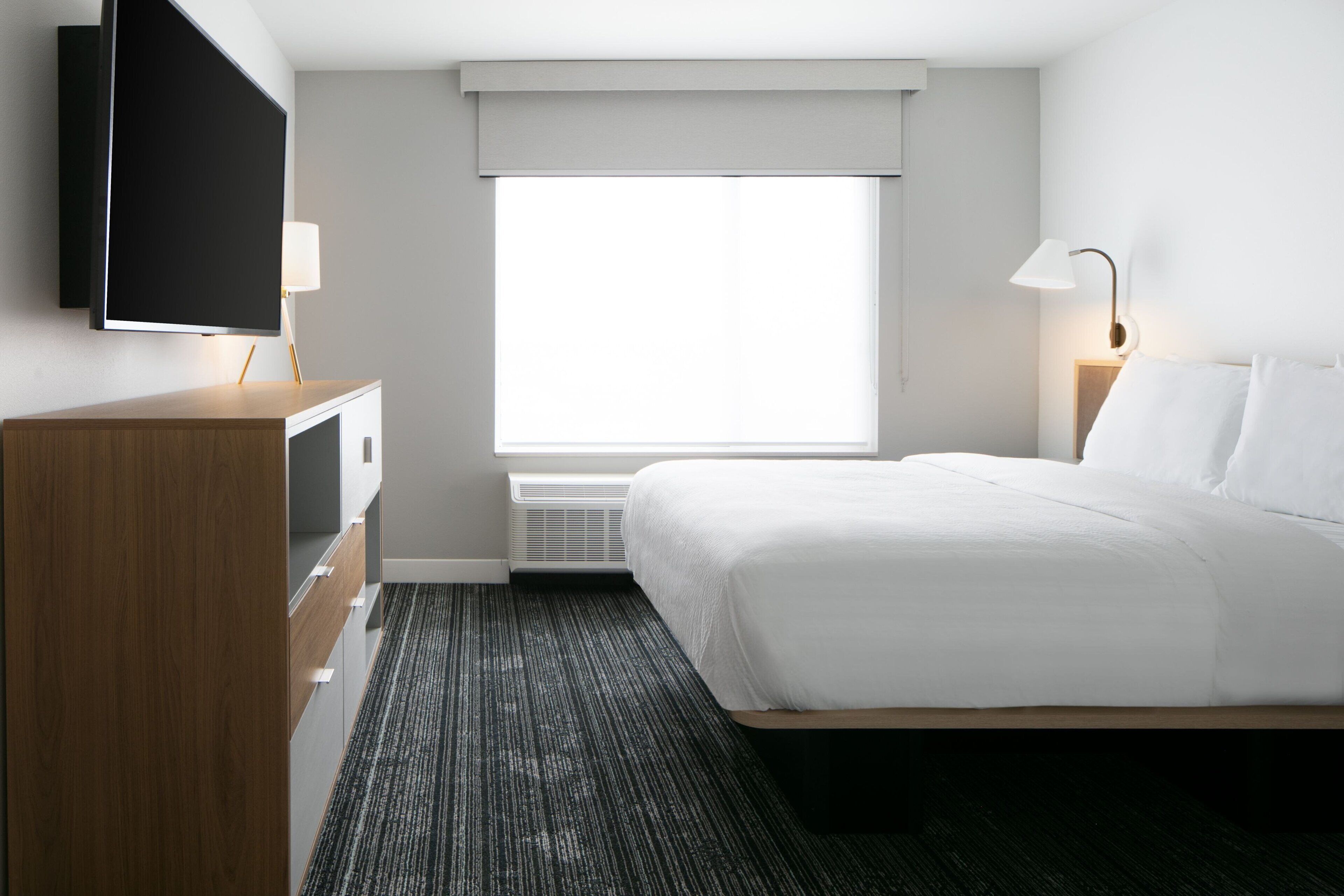 A hotel room with a bed, a TV mounted on the wall, a desk with drawers, a window with blinds, and a bedside lamp.