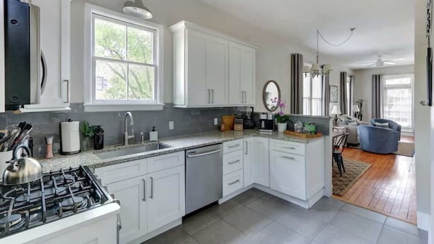A modern kitchen with white cabinetry, a gas stove, a dishwasher, and a dining area with a blue chair.