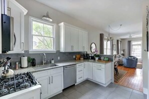 A modern kitchen with white cabinetry, a gas stove, a dishwasher, and a dining area with a blue chair. - The Broad House, Historic | Renewed | Cozy (Savannah)