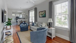 A modern living room with a sofa, armchairs, a coffee table, a TV, a ceiling fan, a dining area, and a kitchen in the background.