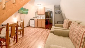 Ett kompakt bostadsutrymme med ett matbord och stolar i trä, en soffa, ett köksområde med skåp och vitvaror samt ett sluttande tak.
