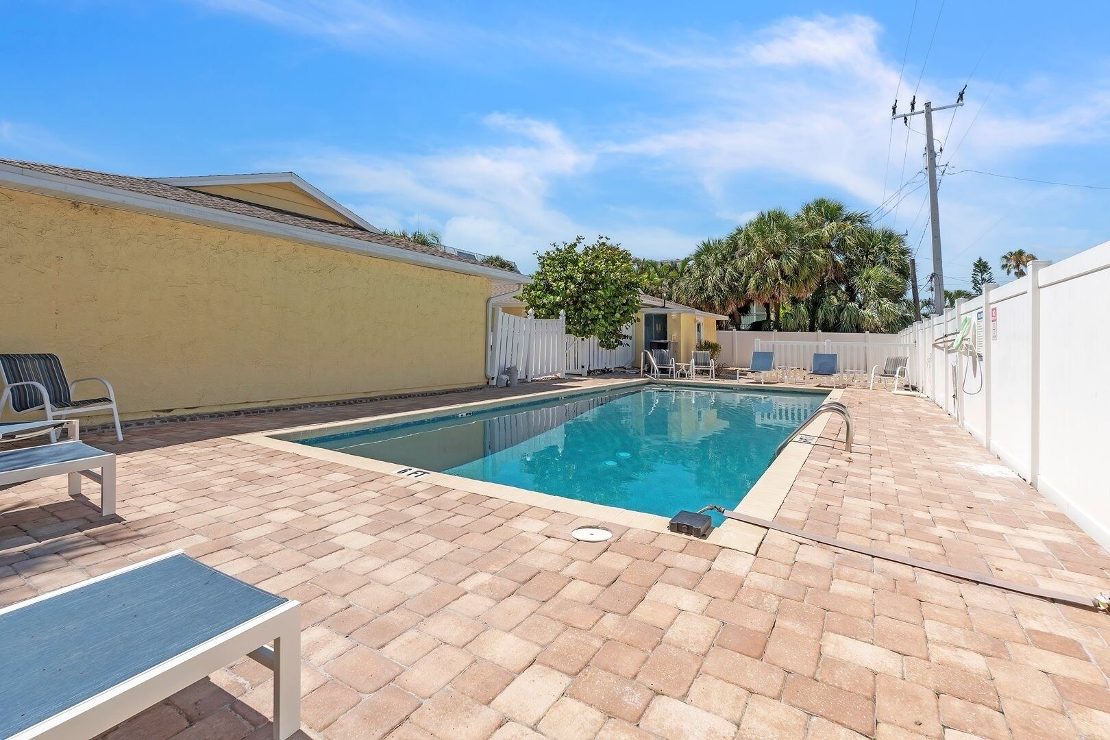 A rectangular swimming pool with a tiled deck, surrounded by a white fence and palm trees.