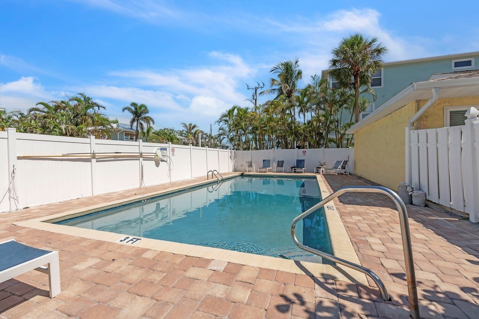A rectangular pool with a metal ladder, surrounded by a white fence and palm trees.