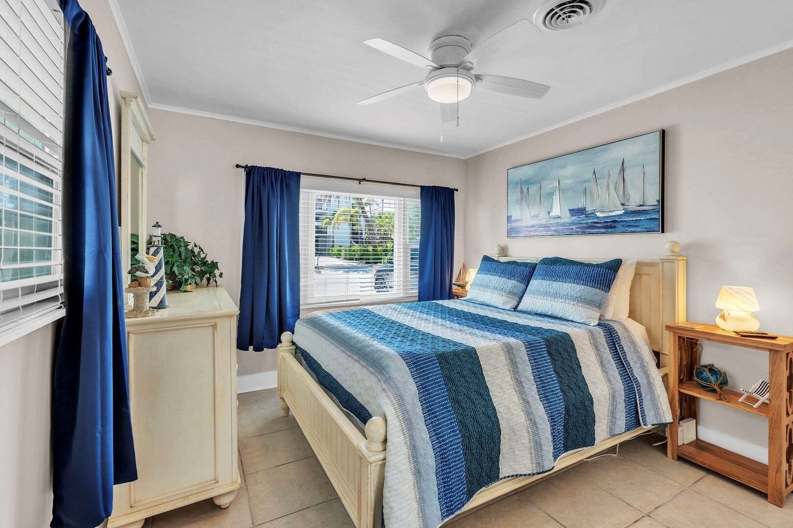 A bedroom with a bed, a nightstand, a ceiling fan, a window with curtains, and a painting on the wall.
