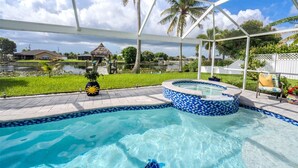 A swimming pool with a hot tub, surrounded by a covered area with palm trees and a view of a lake. - Romantic vacation spot by the water with private heated pool and spa, kayaks! (Cape Coral)
