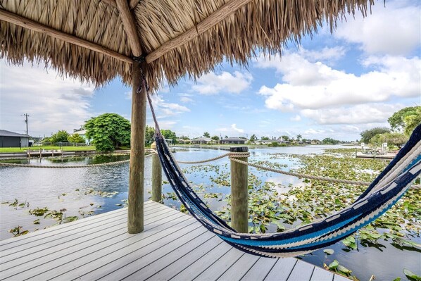 A hammock under a thatched roof, wooden deck, and water with lily pads.