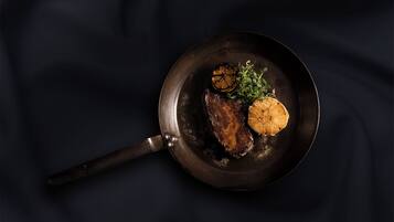 Uma frigideira com um bife, uma fatia de limão e algumas ervas sobre uma superfície escura.