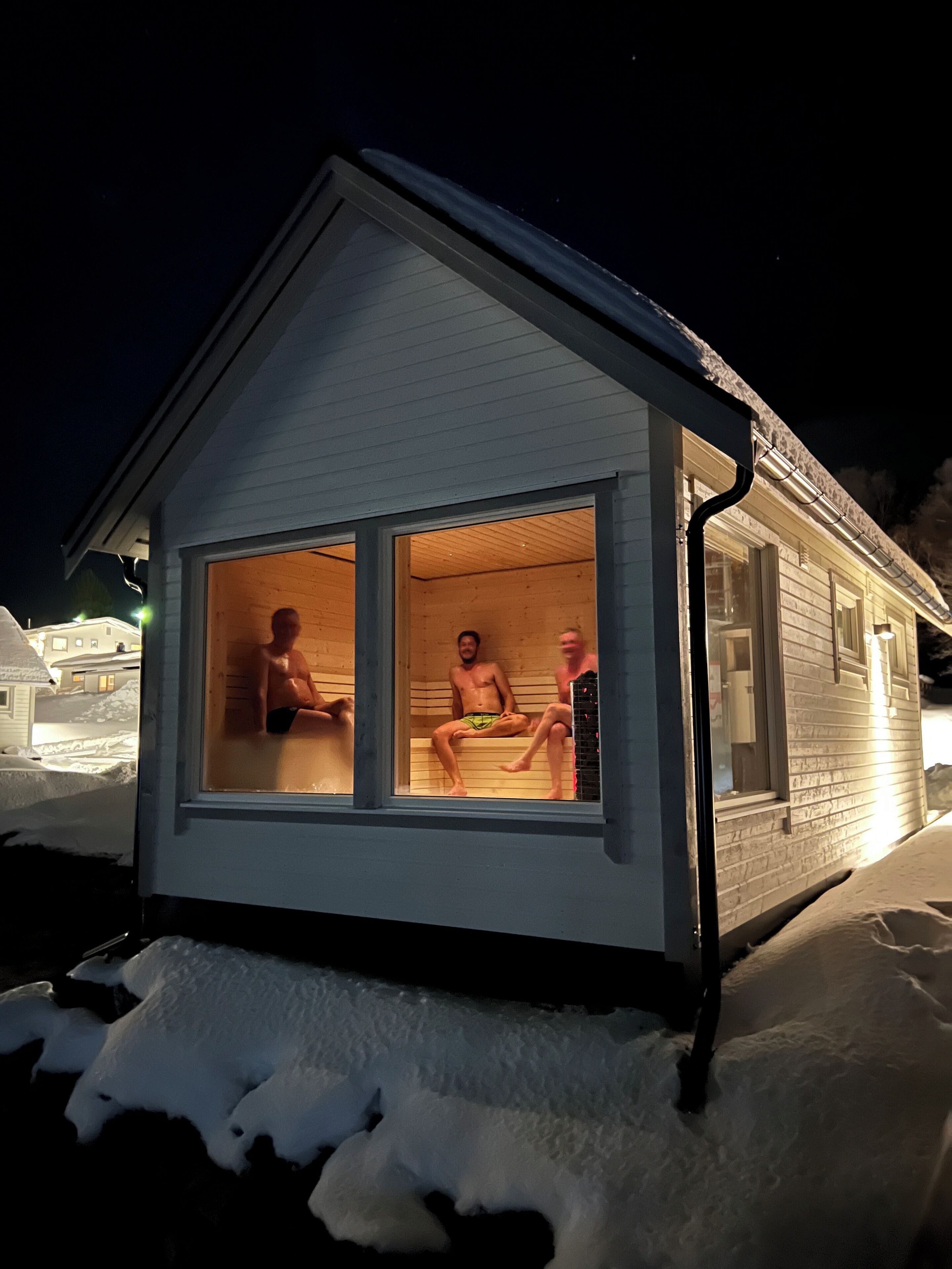 Une petite cabane en bois, d’un seul étage, avec un toit en pente, de grandes fenêtres et un sol recouvert de neige.