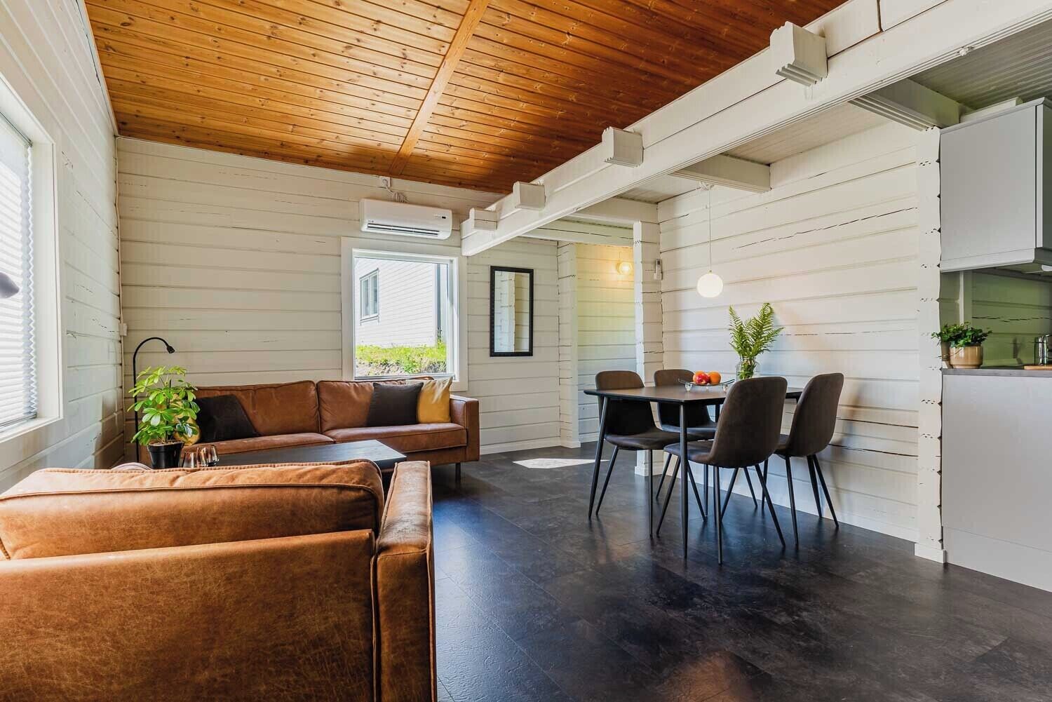 A modern interior with a wooden ceiling, white walls, a leather sofa, a dining table with chairs, and a kitchen area.