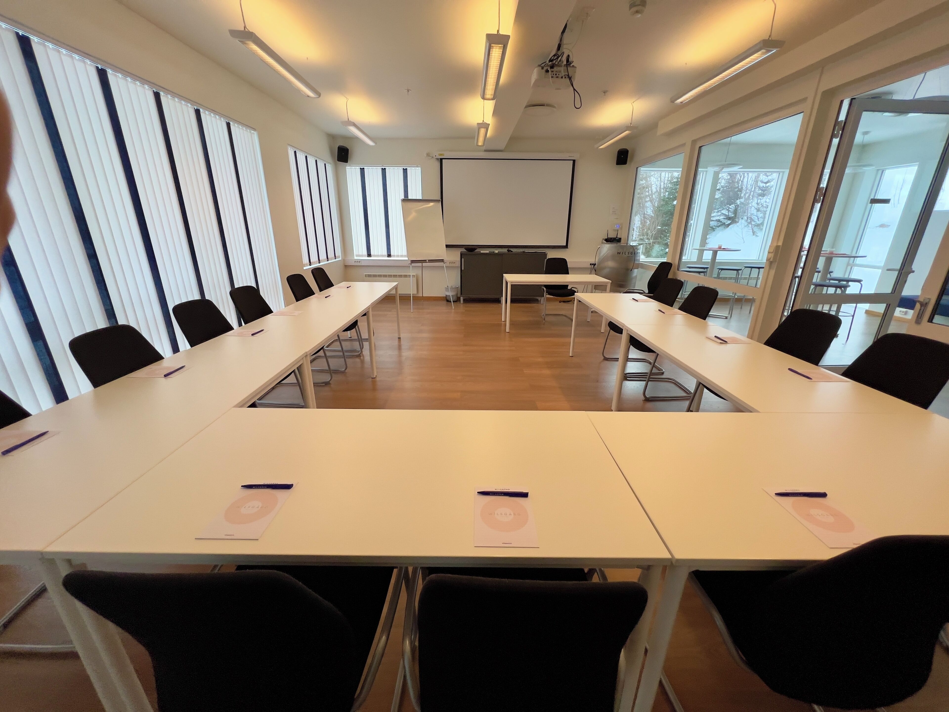 Una sala conferenze con lunghe tavolate, sedie nere, uno schermo per proiettore e ampie finestre.