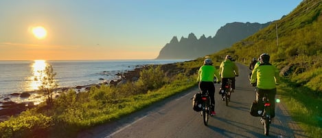 A paved coastal road with cyclists, a sunset, and a mountainous backdrop.