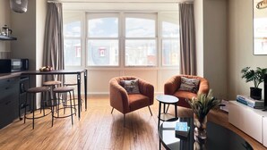 A modern living room with a wooden floor, a black coffee table, two brown armchairs, a small round table, and a large window with curtains. - Villa Adèle, Loft Lumineux et vue Mer - Appt 04 (Mers-les-Bains)