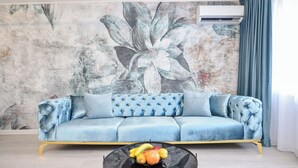 A living room with a blue sofa, a round glass coffee table with fruit, and a floral mural on the wall. - Celeste - One Bedroom Apartment - Next to Victoria Avenue and Cismigiu Park (Bucharest)