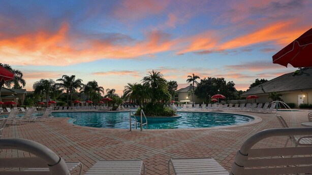Zona de piscina con sillones de descanso y sombrillas, rodeada de palmeras y un cielo al atardecer.