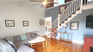 Ein Wohnzimmer mit einem grauen Ecksofa, einem weißen Couchtisch, einem Holzschaukelstuhl und einer Treppe mit weißer Geländer.