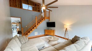 Ein geräumiges Wohnzimmer mit einer großen Eckcouch, einer Holztreppe, einem Flachbildfernseher und einem Deckenventilator.