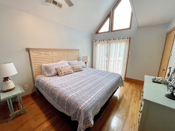 A bedroom with a wooden bed, a nightstand with a lamp, a window with curtains, and a wooden floor.