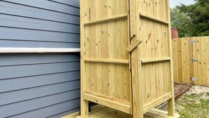 Une structure en bois avec un toit incliné, qui pourrait être un abri de jardin ou un local de rangement, située à côté d’une maison dont le revêtement extérieur est bleu.