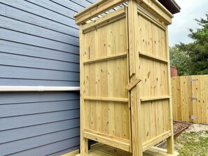 Une structure en bois avec un toit incliné, qui pourrait être un abri de jardin ou un local de rangement, située à côté d’une maison dont le revêtement extérieur est bleu.
