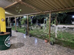 Un espace extérieur aménagé en patio, avec un sol carrelé, des plantes en pot et une partie couverte dotée d’un pilier jaune.
