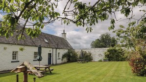 A white building with a sloped roof, a stone wall, and a grassy yard with picnic tables and bird feeders. - Fordie South Cottage (Crieff)