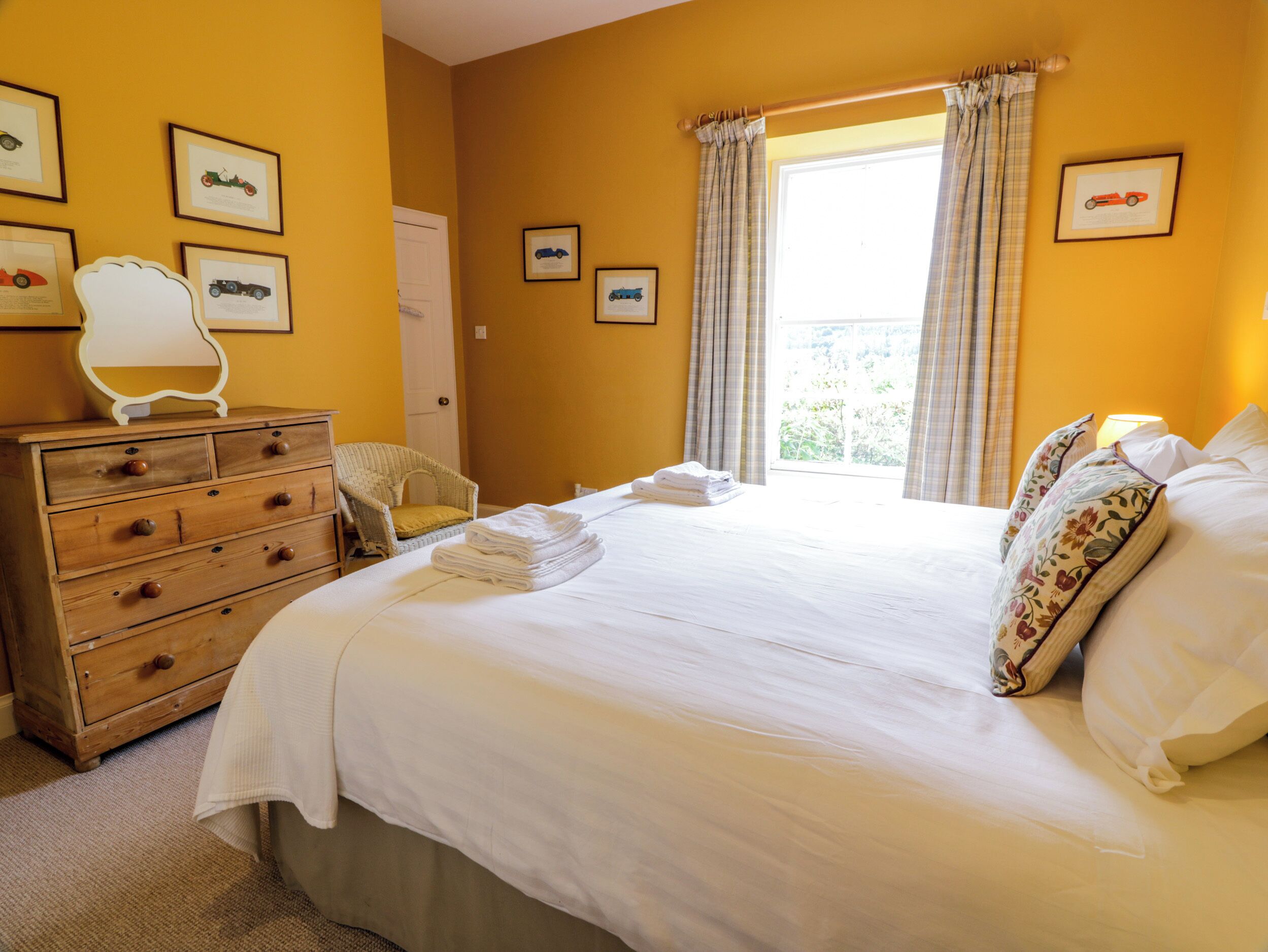 Habitación de hotel con una cama grande, cómoda de madera y una silla. Se observan cuadros enmarcados en la pared y una ventana con cortinas.