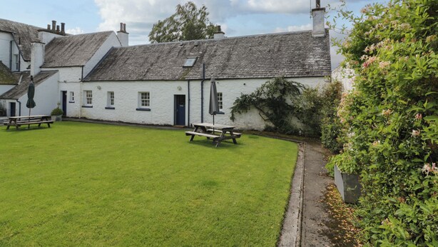 A traditional white building with a grassy courtyard, two picnic tables, and a small garden area with flowering plants. - Fordie South Cottage (Crieff)