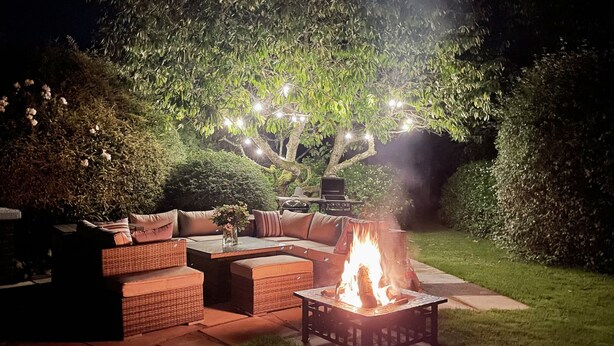 Un espace extérieur aménagé en terrasse, avec un foyer, des meubles en osier et des guirlandes lumineuses suspendues à un arbre.