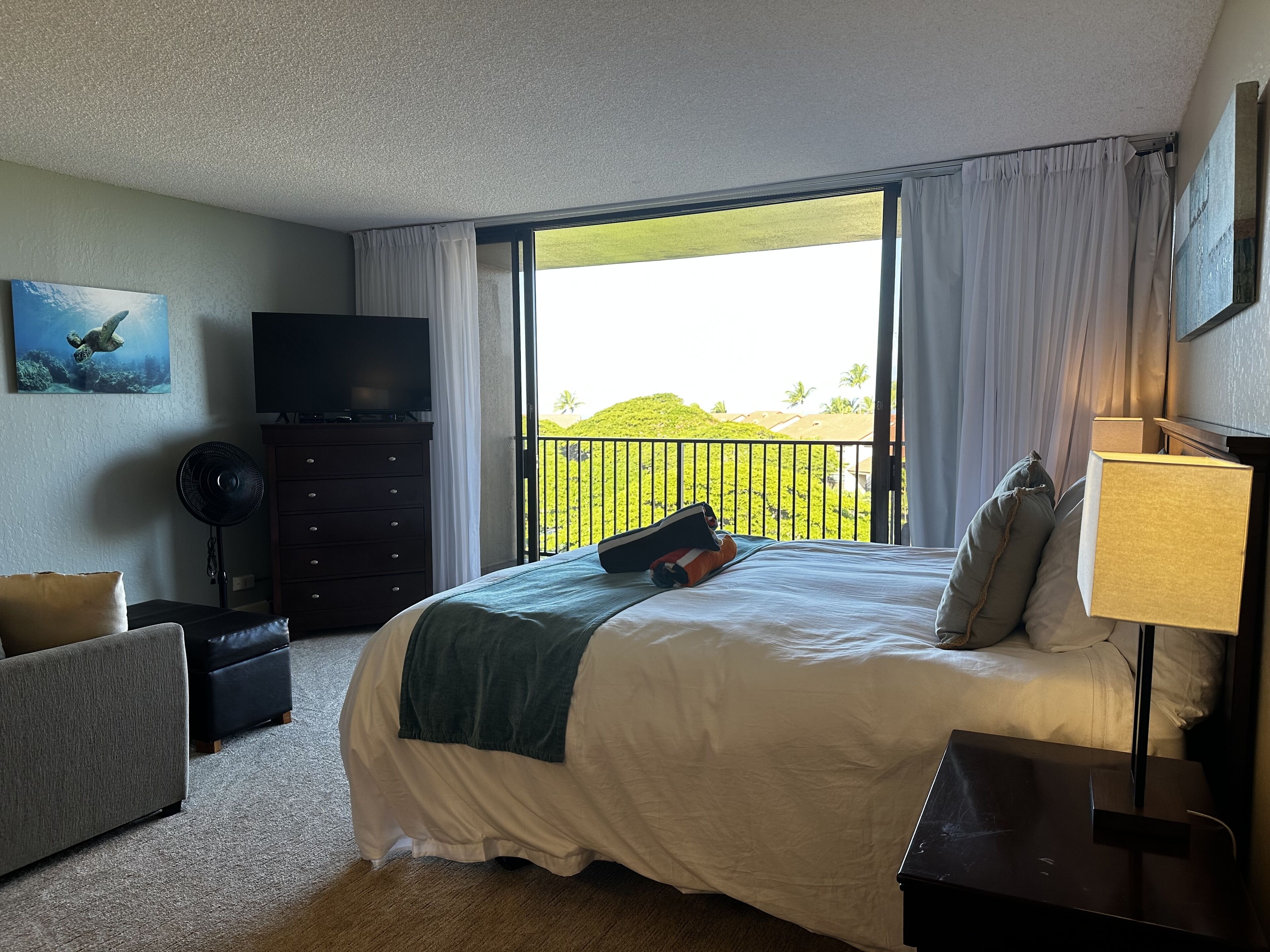 Habitación de hotel con una cama grande, televisión, escritorio, silla y balcón con vistas a zonas verdes.