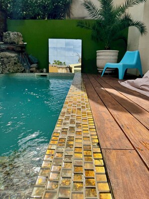 Une piscine avec un bord carrelé, une terrasse en bois, une chaise bleue et une plante en pot.