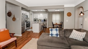 A modern living room with a grey sofa, a coffee table, and a dining area with a table and chairs. A kitchen with white cabinets and stainless steel appliances is visible in the background. - 3BR Near Cleveland & CVNP | Sunroom + Ping-Pong (Independence)
