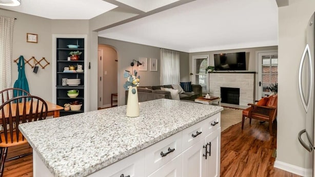 A modern kitchen with granite countertops, white cabinetry, and a central island. There is a dining area with a wooden table and chairs, and a living room with a fireplace and seating. - 3BR Near Cleveland & CVNP | Sunroom + Ping-Pong (Independence)
