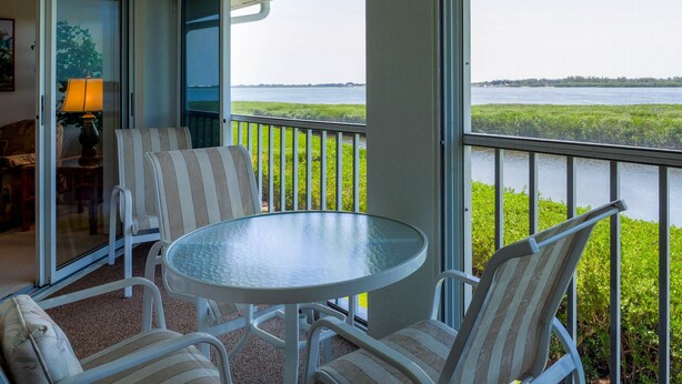 A balcony with a glass table and striped chairs, overlooking a body of water and green vegetation. - Perico Bay Club · 30-Night Min · Intracoastal (Bradenton)